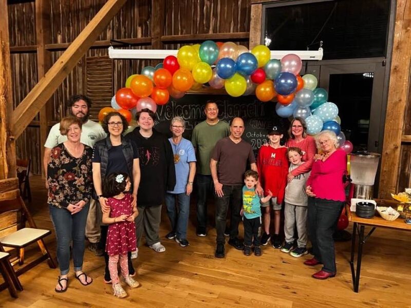 A group of people, including children, stands smiling under a colorful balloon arch in a wooden indoor setting with party decorations.