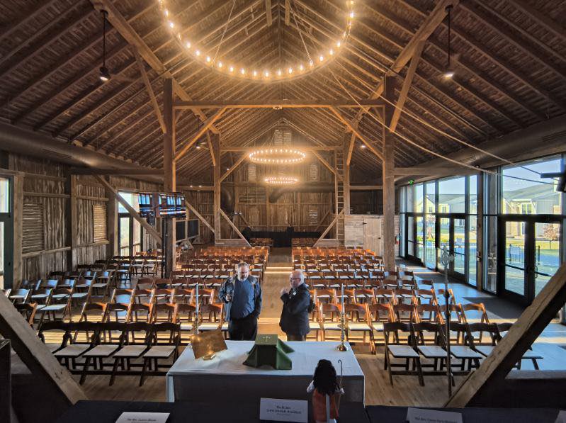 A rustic venue with wooden beams, empty chairs, and two people near a table. Large windows reveal an outdoor setting.