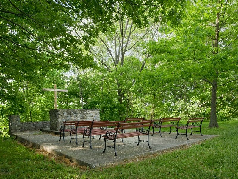 outdoor altar A serene outdoor chapel with wooden benches and a stone altar featuring a cross, surrounded by lush green trees.