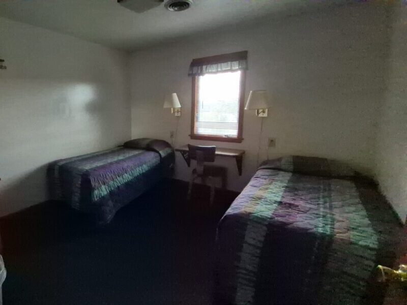 A beige house with a wooden deck is surrounded by greenery and a brick pathway, nestled in a wooded area.