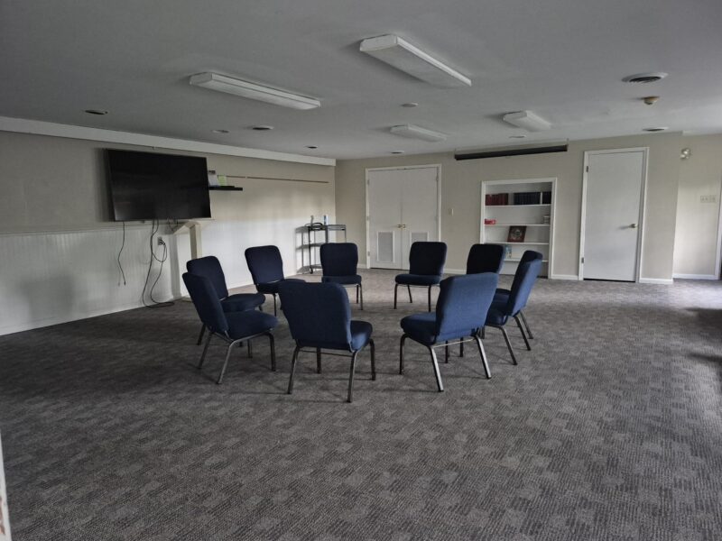 Modern two-story building with beige siding and stone accents, surrounded by a lawn and trees. Foreground features outdoor seating and paving.
