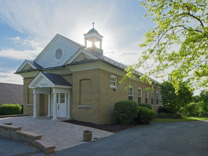 Claggett-20 A small brick church with a steeple is surrounded by lush greenery, under a partly cloudy sky.