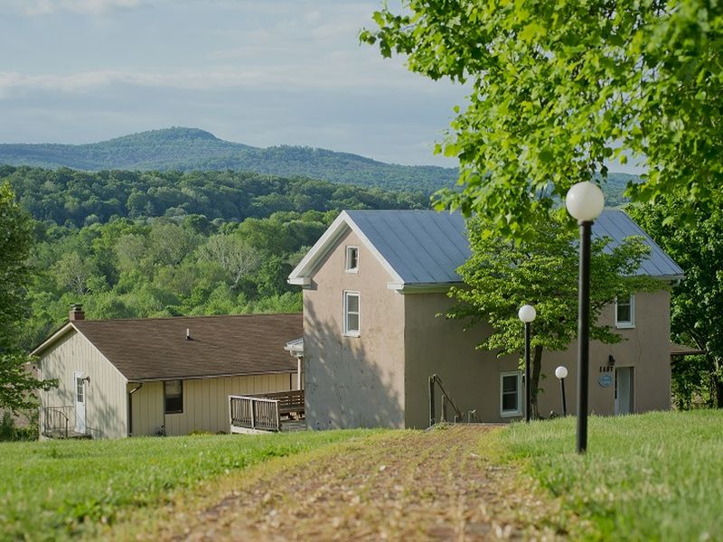Two houses nestled in lush green countryside, with rolling hills in the background. A dirt path leads past street lamps.