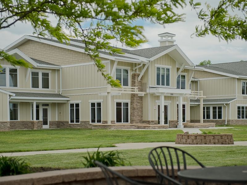 A large, modern two-story building with beige paneling stands amidst green lawns, set against a cloudy sky. Metal chairs feature in the foreground.