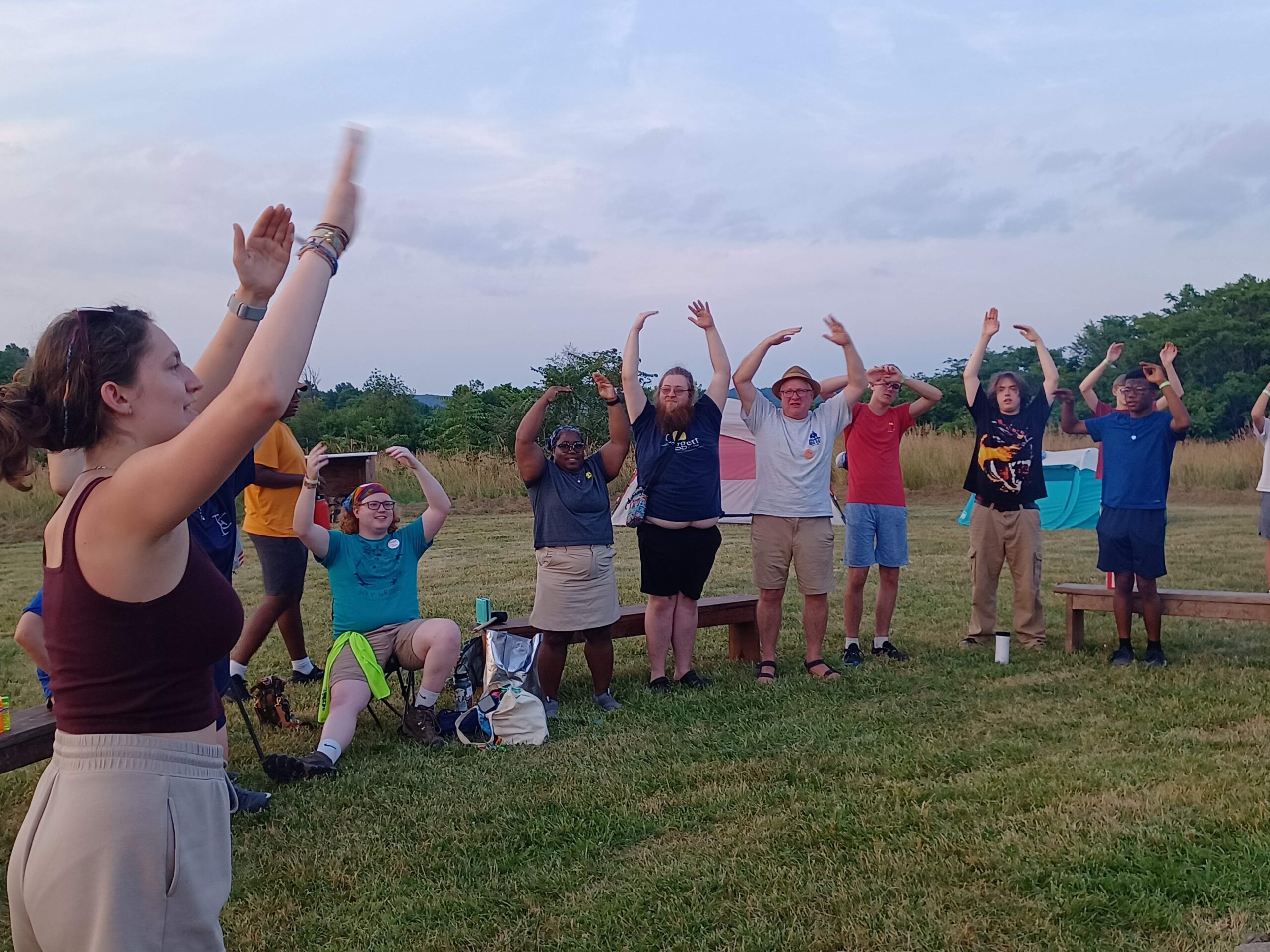 Group Exercise A group of people outdoors is doing a group exercise with arms raised, surrounded by grassy fields and tents in the background.