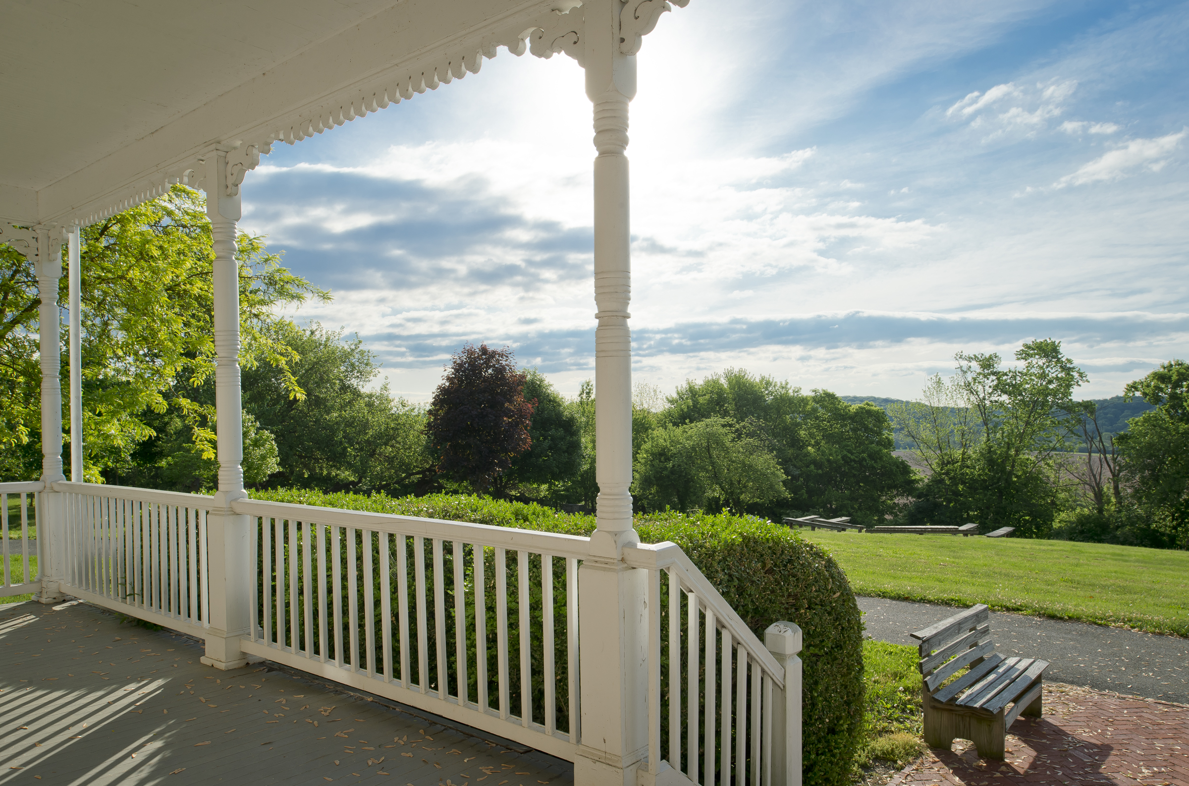 A scenic porch view features lush greenery, a wooden bench, and partly cloudy skies, creating a peaceful and inviting outdoor atmosphere.