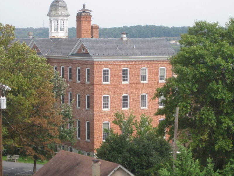 Historic brick building with cupola and chimney, surrounded by lush trees. The scene conveys a serene, timeless atmosphere, blending architecture with nature.
