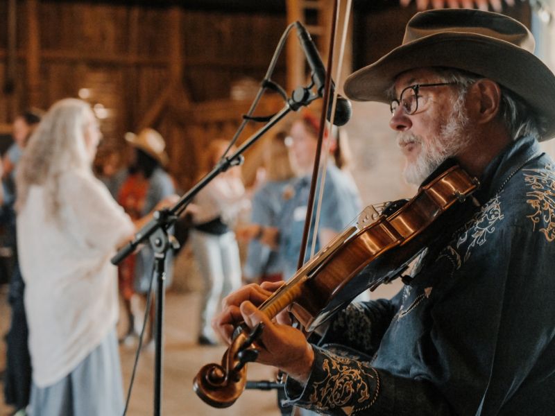 Fiddle Player A person plays the violin in a rustic, indoor setting with others dancing or socializing in the background, creating a lively atmosphere.