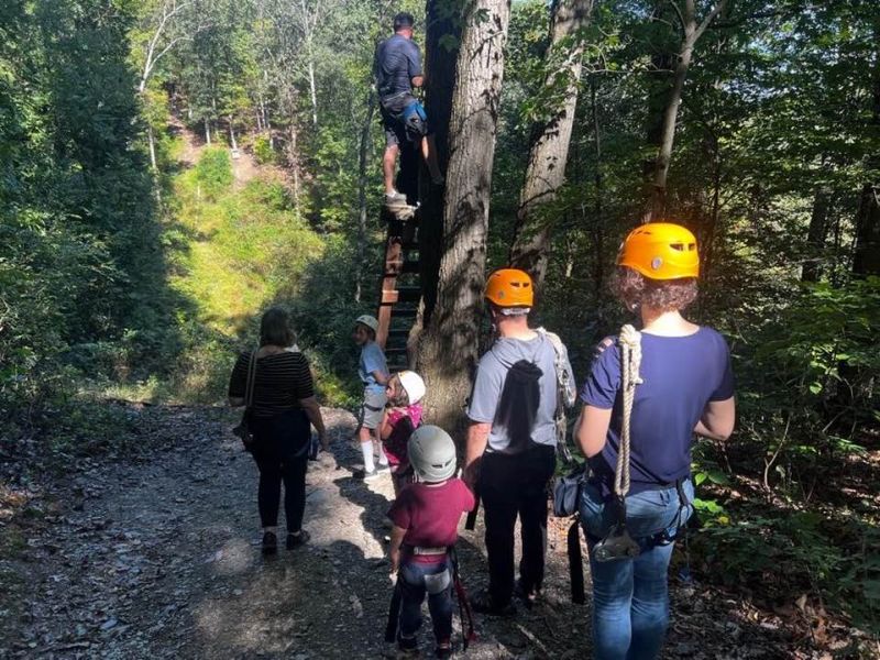 Hike-Trail A group, including children, ready for zip-lining in a forest; wearing helmets and safety gear near a tree platform. No landmarks visible.