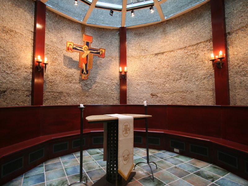 Thomas Beckett Chapel A small chapel interior with stone walls features a crucifix, altar, and lit candles, creating a serene, reverent atmosphere.