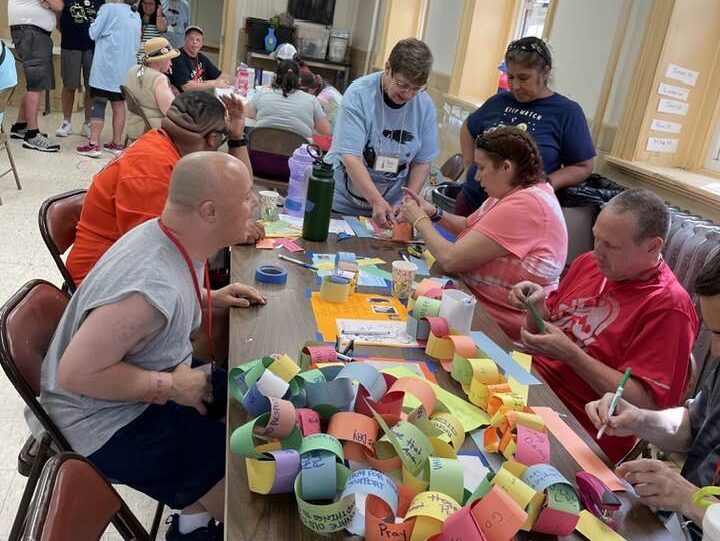 adult camp art time People gather around a table creating colorful paper chains in a room with brown chairs and fluorescent lighting.