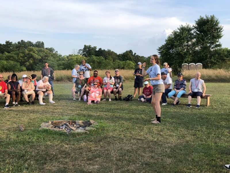 adult camp fireside A group of people gathers outdoors near a small campfire pit, surrounded by trees, under a clear sky with grassy fields.