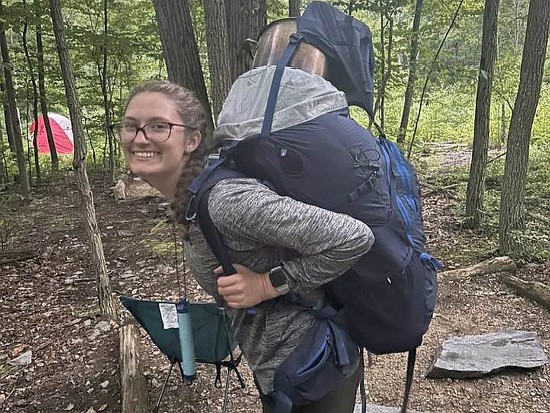 backpacking A person wearing a gray shirt carries a large backpack in a wooded area with a red tent in the background.