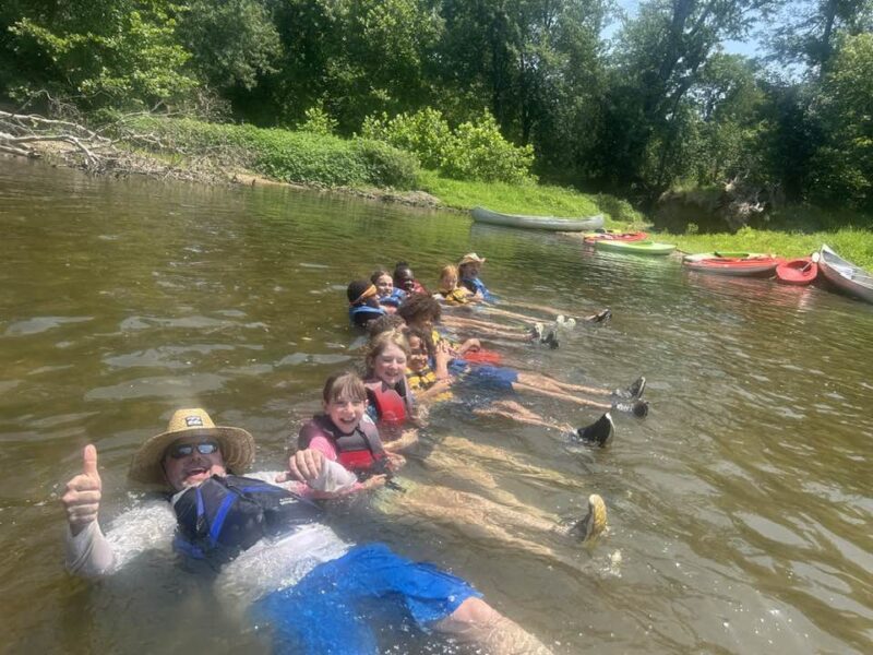 campers in the lake A group of people and children relax in a river, wearing life vests, with kayaks nearby. They seem to be enjoying sunny weather.