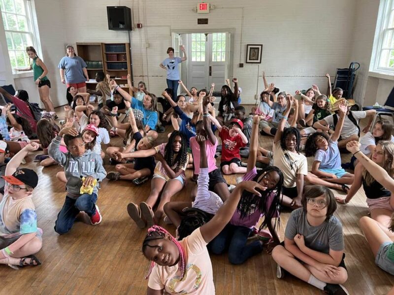 campers inside stretching A large group of children and a few adults sit on a wooden floor, raising their hands, in a brightly lit room.