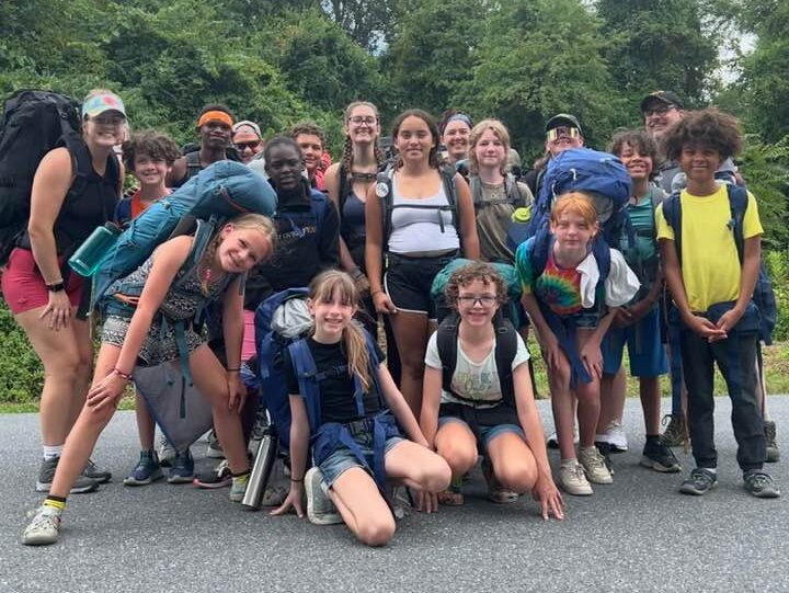 campers on a hike A group of people and children with backpacks gather on a road, surrounded by lush green trees and a partly cloudy sky.