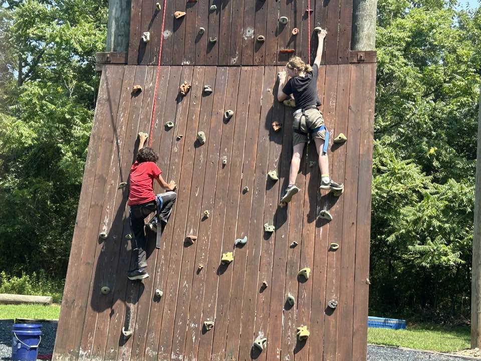 campers on rock wall Two people climb a wooden outdoor rock wall surrounded by greenery, with climbing gear and harnesses visible, under a clear sky.