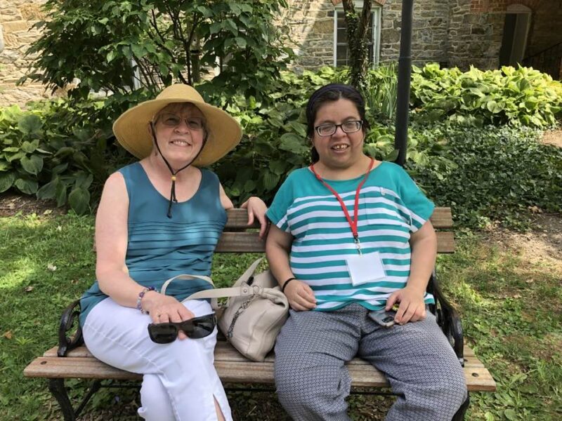 Two people sitting on a bench outside; one wearing a straw hat. Background features a stone building and lush greenery.