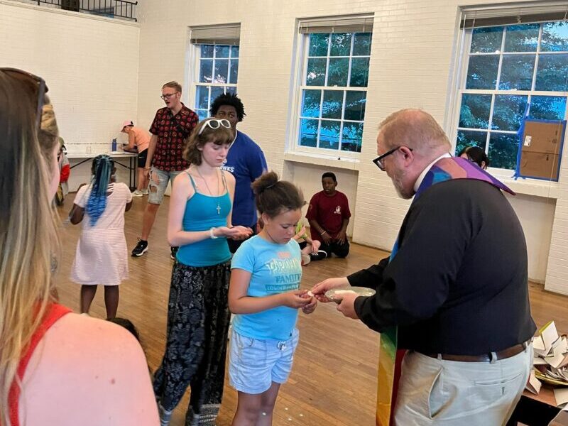 chaplain-communion A person in clergy attire interacts with others in a hall, featuring high ceilings, wooden floors, and large windows showing greenery outside.