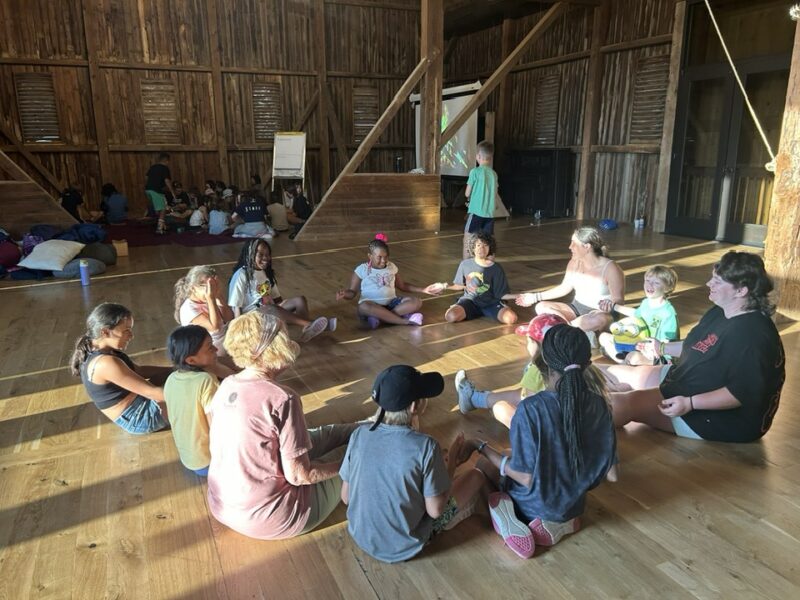 circle time at camp A group of children and two adults sit in a circle inside a rustic wooden barn, engaging in a playful activity together.