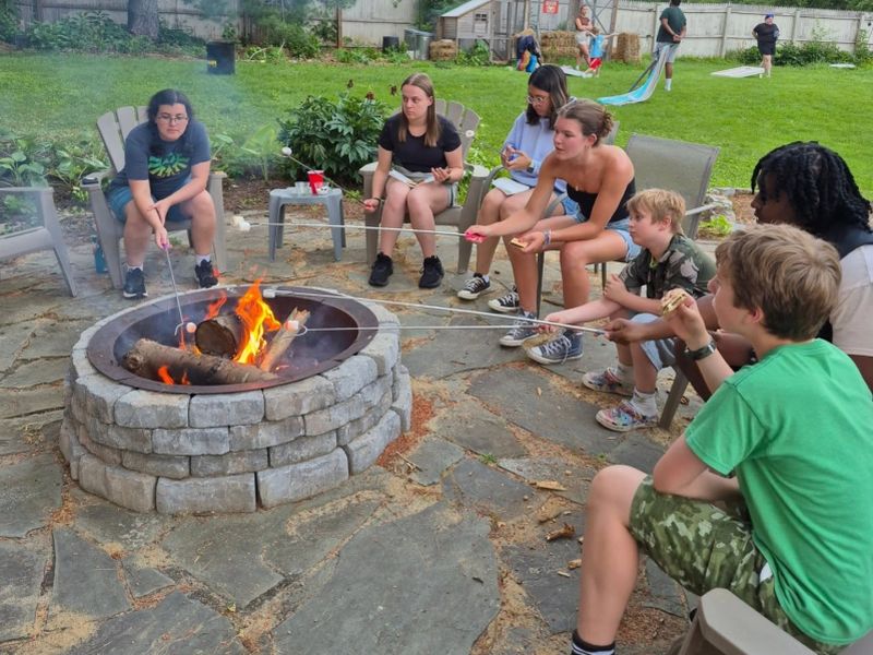 Several people sitting around an outdoor fire pit, roasting marshmallows, surrounded by greenery and patio stones in a backyard setting.