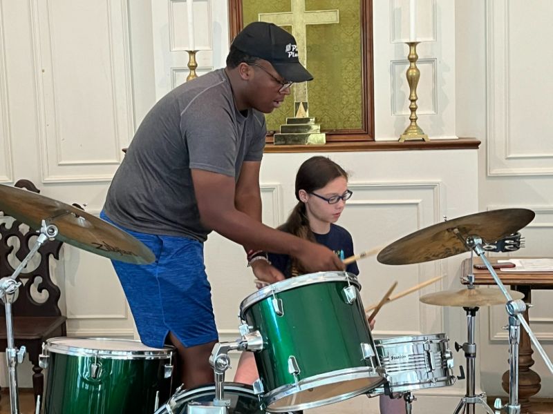 drum lesson A person assists a child learning to play a drum set in a room with a decorative cross and candlesticks.