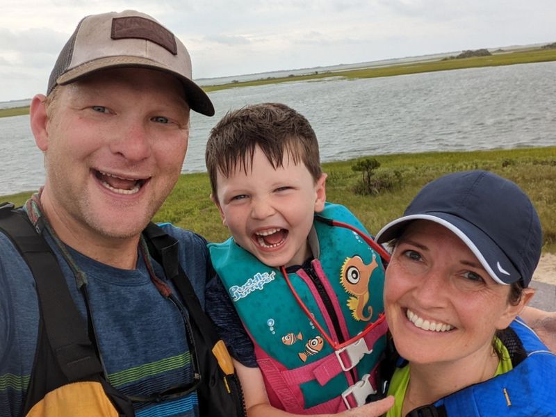 family by the water Three people smiling near a body of water, with grassy landscape in the background. Two adults and one child, wearing outdoor gear.