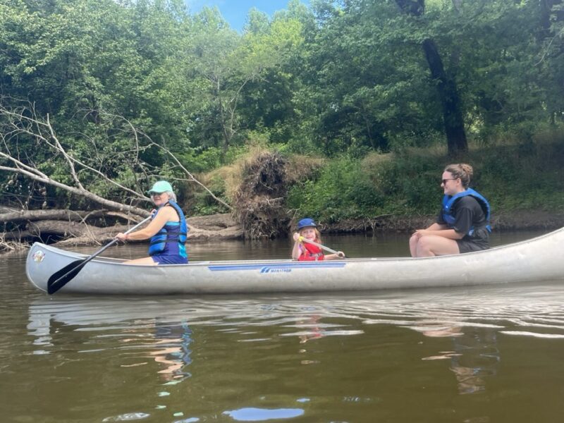 family kayak trip Three people in a canoe paddling on a calm river, surrounded by lush green trees and natural scenery.