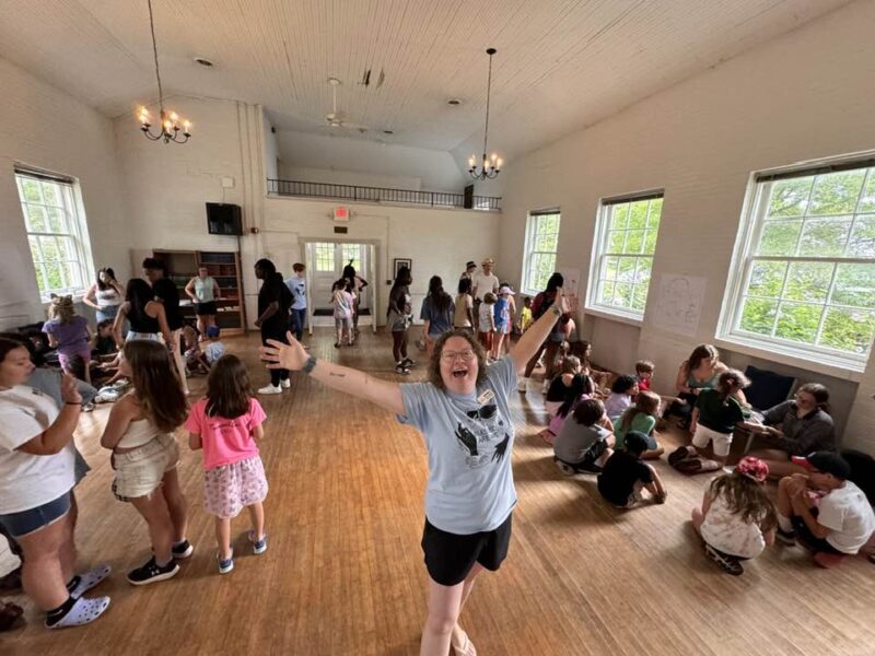 fun at camp A cheerful person stands in a bright room with large windows, surrounded by groups of children and adults engaging in various activities.