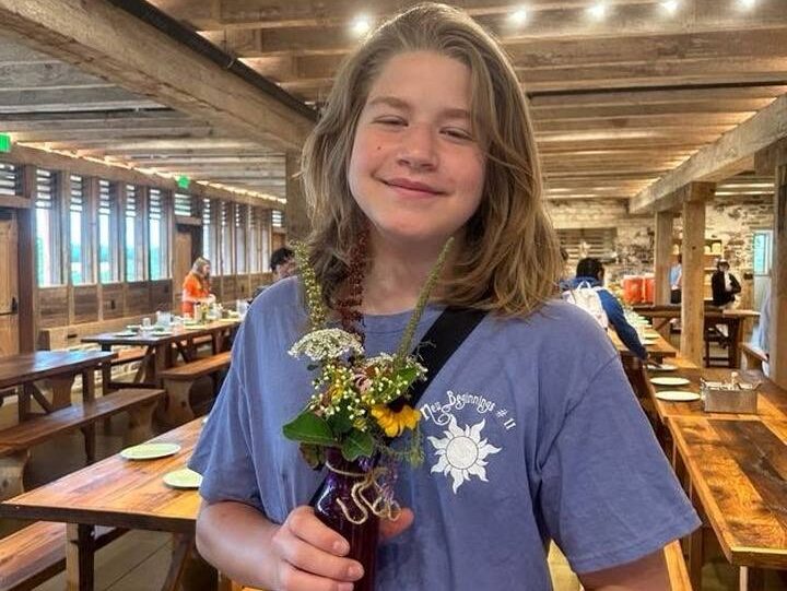 teen with flowers A smiling person holds a small vase with flowers in a rustic wooden interior, featuring long tables and warm lighting.