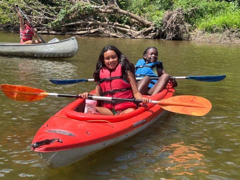 Two kids enjoying a sunny day kayaking in a red kayak on a calm river, surrounded by lush greenery.