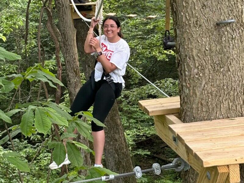 girls on ropes course Two people engage in a forest adventure activity, navigating ropes among trees on an elevated obstacle course. Lush greenery surrounds the scene.