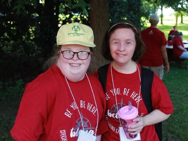 Two people wearing red shirts and lanyards smile under a tree. Others gather in the background at an outdoor event.