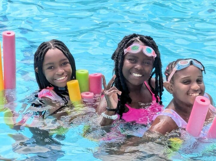 girls swimming Three children in a pool, smiling and holding colorful pool noodles, wearing swimsuits and goggles, surrounded by bright blue water.