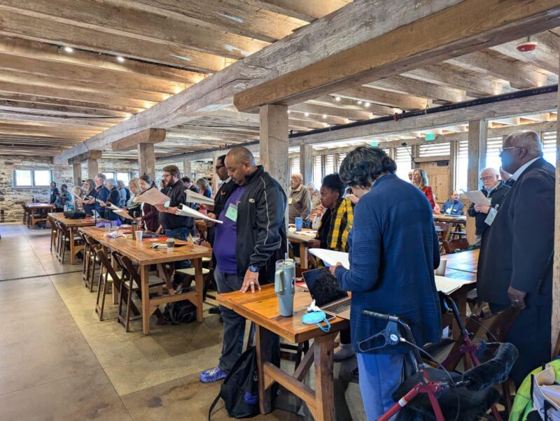 group in barn at session A group of people stand inside a rustic hall with wooden beams, reading from papers at wooden tables.