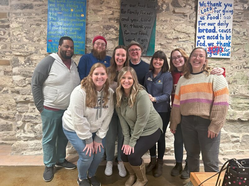 group of volunteers Nine people posing happily inside a rustic stone-walled room with colorful signs, likely for a gathering or event. No recognizable landmarks present.