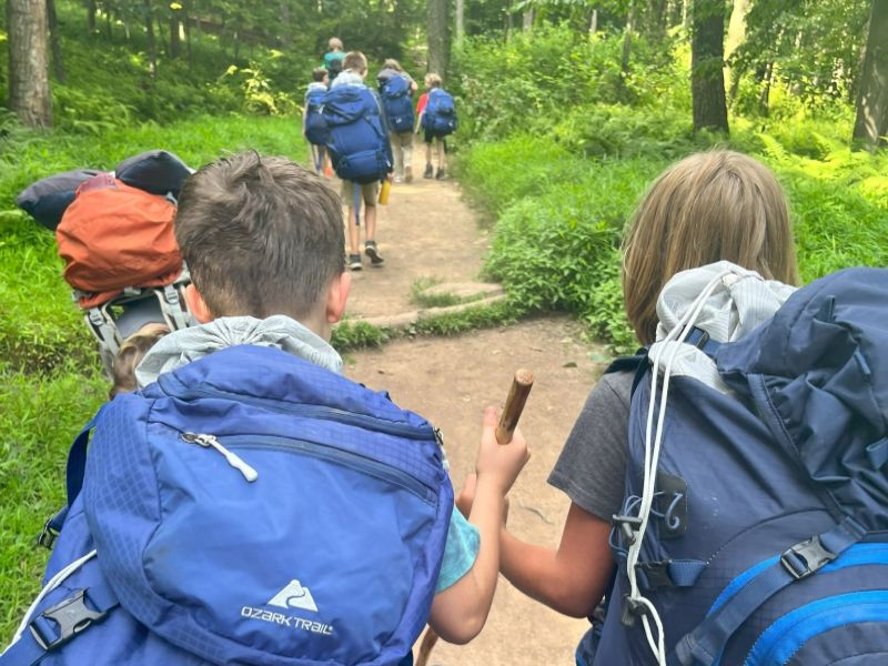 hiking trail A group of backpackers, including children, hike through a lush forest trail. Two individuals carry large packs and walk confidently.