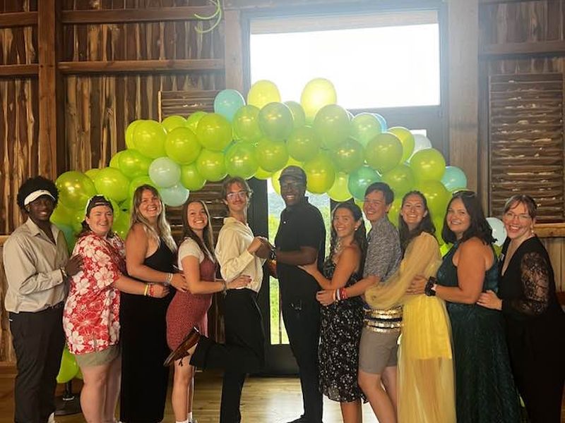 indoor dance party A group of eleven people posing indoors under a green balloon arch, smiling cheerfully in a wooden venue.