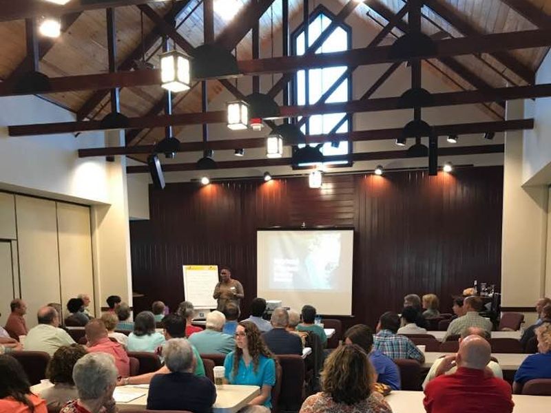 A group of people sits in a wooden-ceiling conference room, listening to a presentation on a screen at the front.