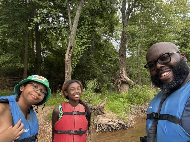 kayaking crew Three people wearing life jackets smile near a wooded riverbank. Tall trees and lush greenery surround them on a cloudy day.