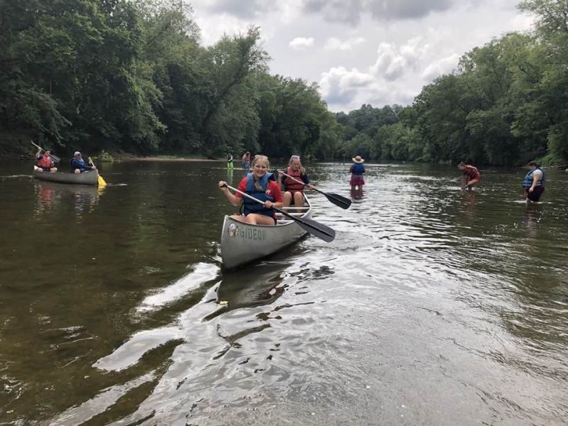 kayaking A group of people is canoeing and wading in a shallow, tree-lined river under a cloudy sky, enjoying an outdoor adventure.
