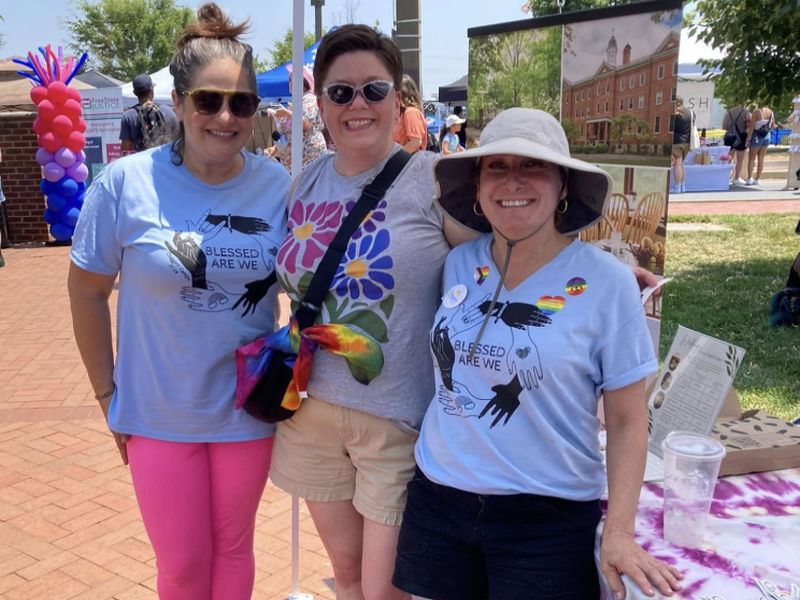 ladies smiling outside Three people smiling at an outdoor event with balloons and a historic brick building in the background, featuring a "Blessed Are We" poster.