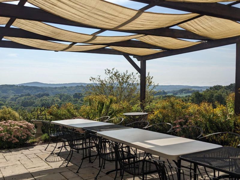 lunch on patio A scenic outdoor patio with tables and chairs overlooks a lush, rolling landscape under a wooden pergola with fabric shades.