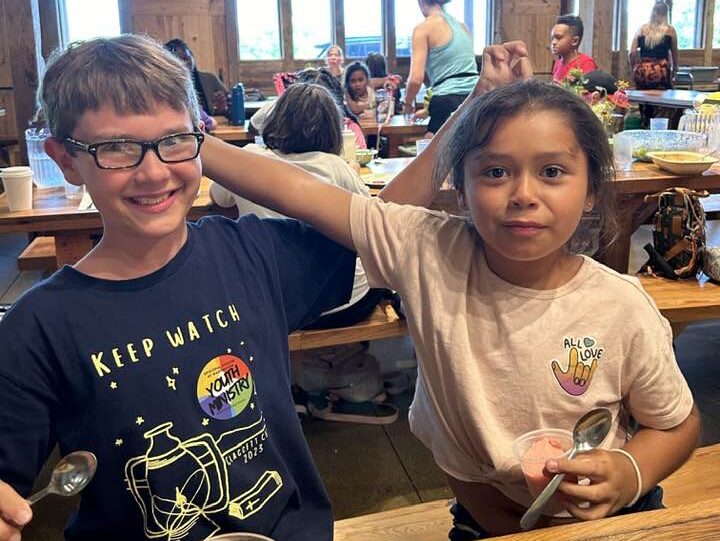 lunch time fun Two children smiling, sitting at a wooden table in a busy dining area, with people and plates of food around. Indoor rustic setting.
