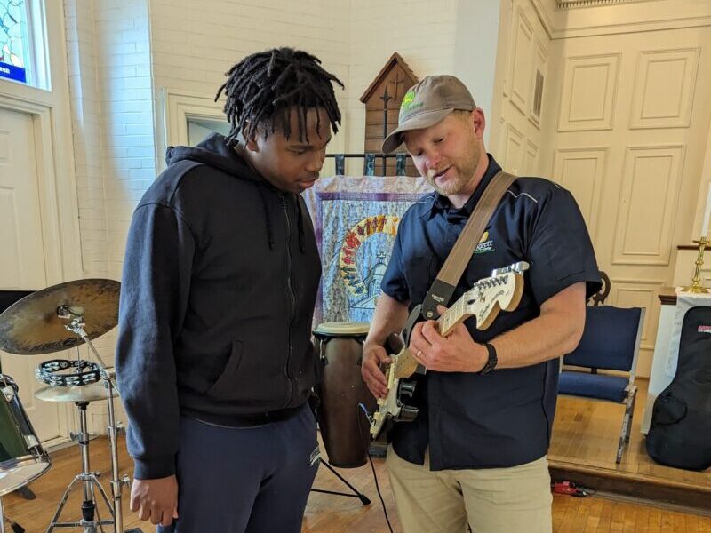 music volunteer Two people in a room with musical instruments; one plays an electric guitar, as they appear to be teaching or learning.