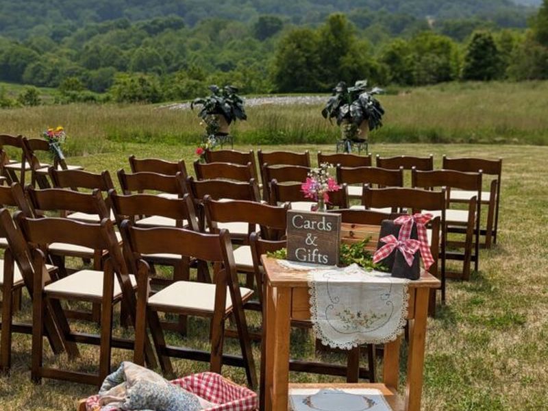 Outdoor wedding setup with wooden chairs and a "Cards & Gifts" table, against a backdrop of lush greenery and distant hills.