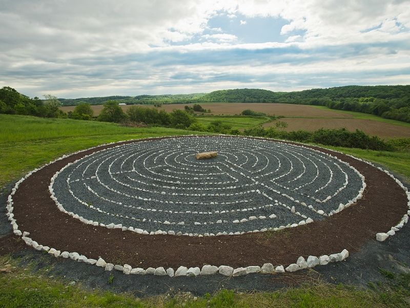 outdoor-labyrinth A circular stone labyrinth set in a grassy field, surrounded by lush countryside and rolling hills under a partly cloudy sky.