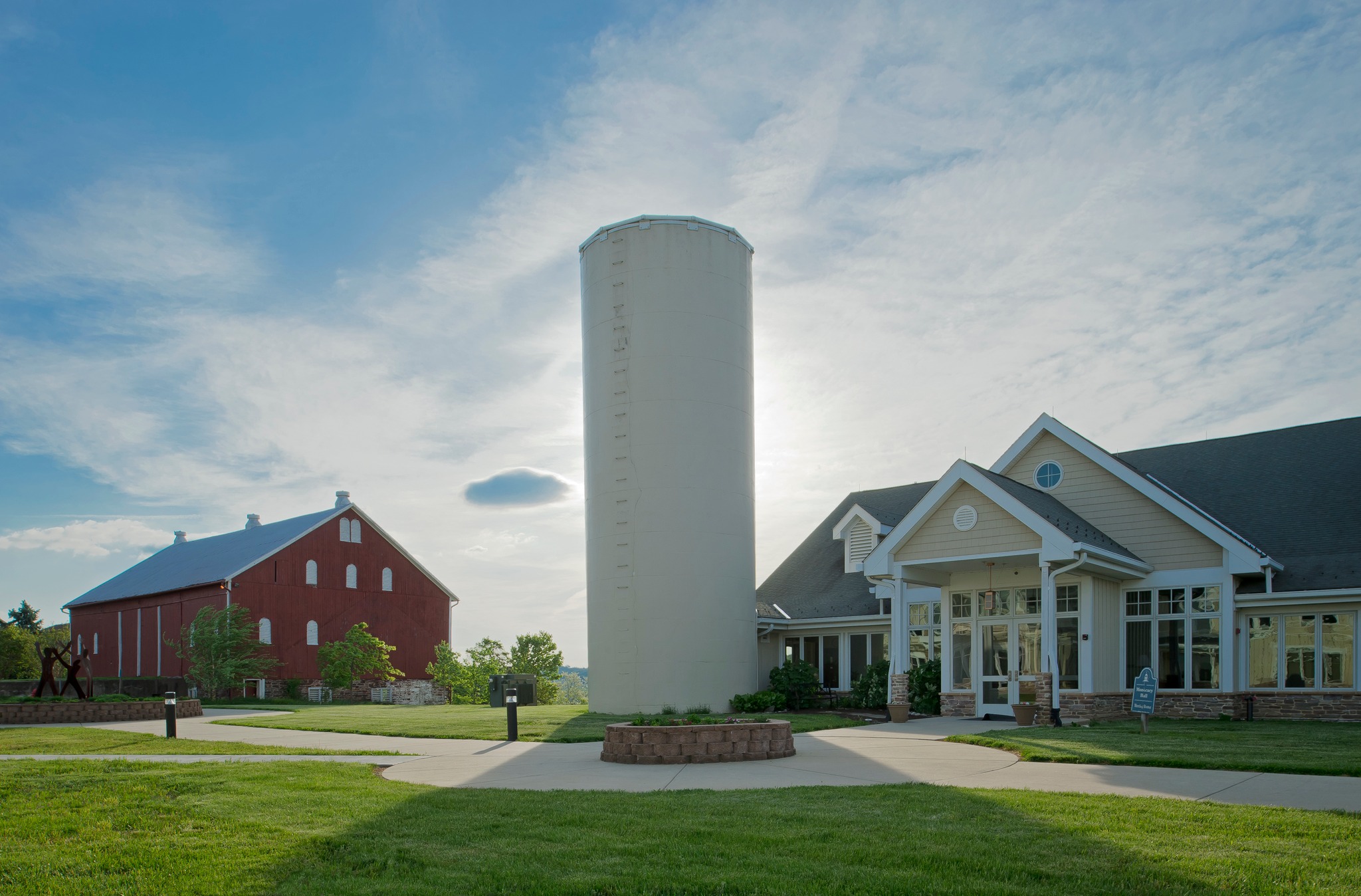 A red barn, large silo, and modern building stand under a partly cloudy blue sky, surrounded by green grass and pathways.