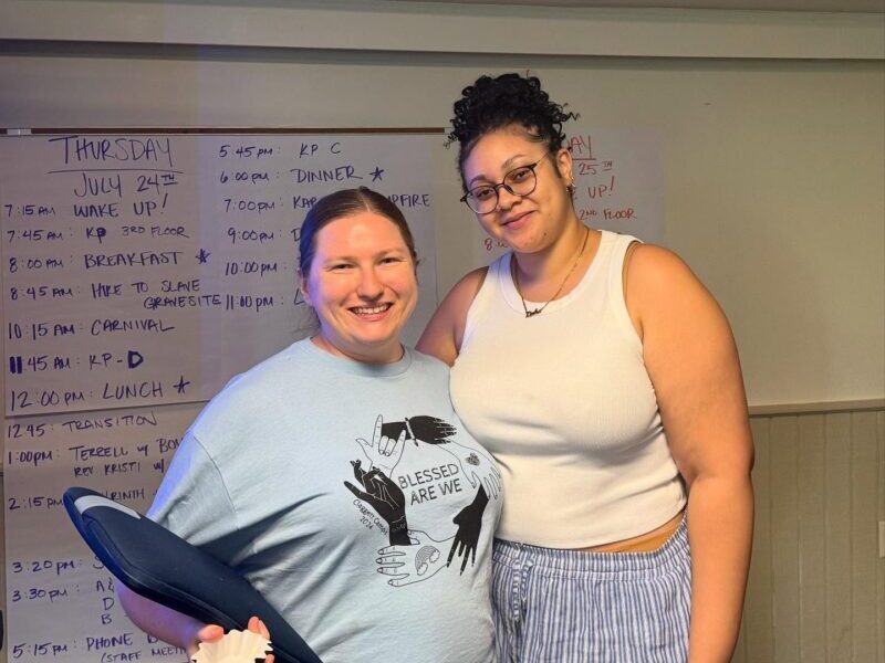 photo of two camp volunteers Two people smile in front of a schedule board. One holds a flip-flop. The schedule lists various activities for Thursday, July 24th.