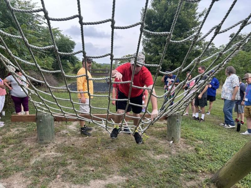 ropes course 2 A person navigates a rope obstacle course outdoors, surrounded by onlookers, amidst green foliage and a partly cloudy sky.